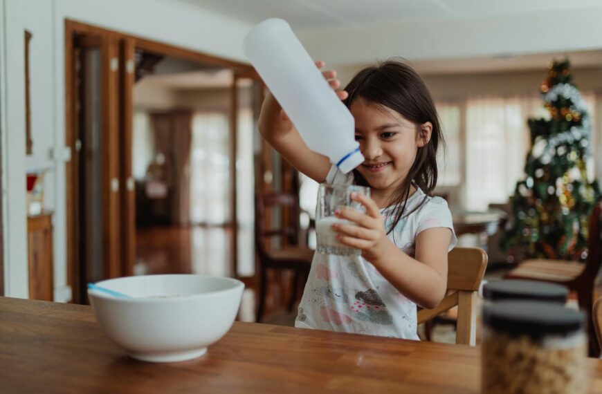 Young girl pouring milk into a glass beside a bowl, enjoying breakfast in a cozy dining room.