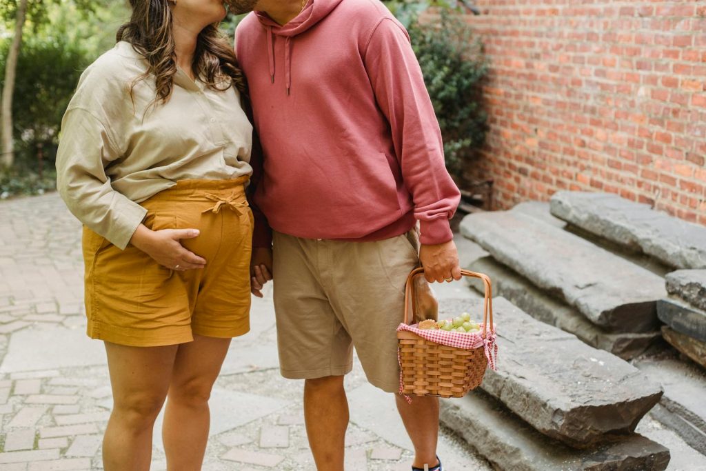 Unrecognizable pregnant woman touching belly and husband with wicker basket caressing each other while standing on veranda near brick building
