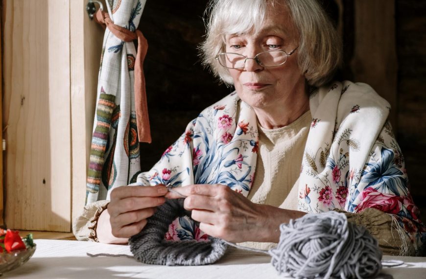 Senior woman knitting with gray yarn near a sunlit window, showcasing peaceful retirement activities.