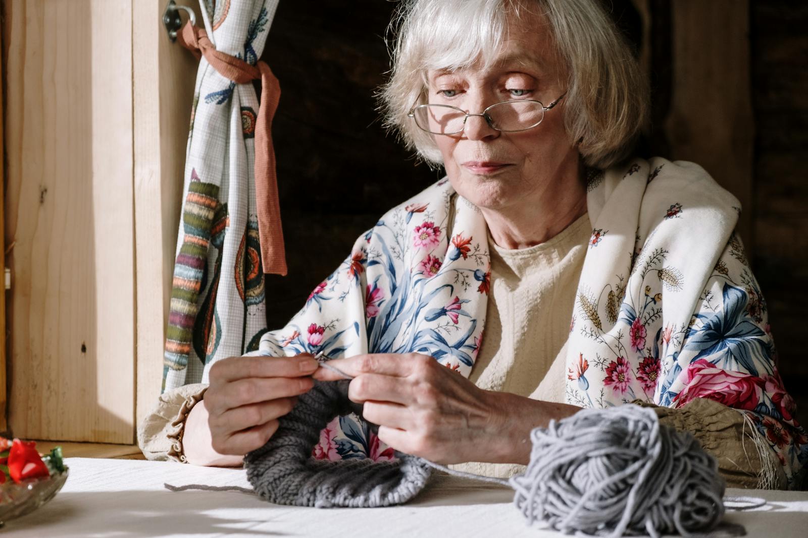 Senior woman knitting with gray yarn near a sunlit window, showcasing peaceful retirement activities.