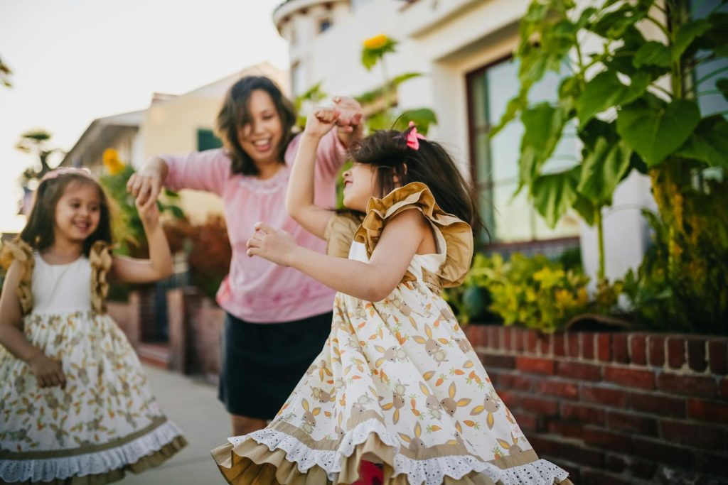 A mother dances with her twin daughters in a lively outdoor setting, enjoying a sunny day.