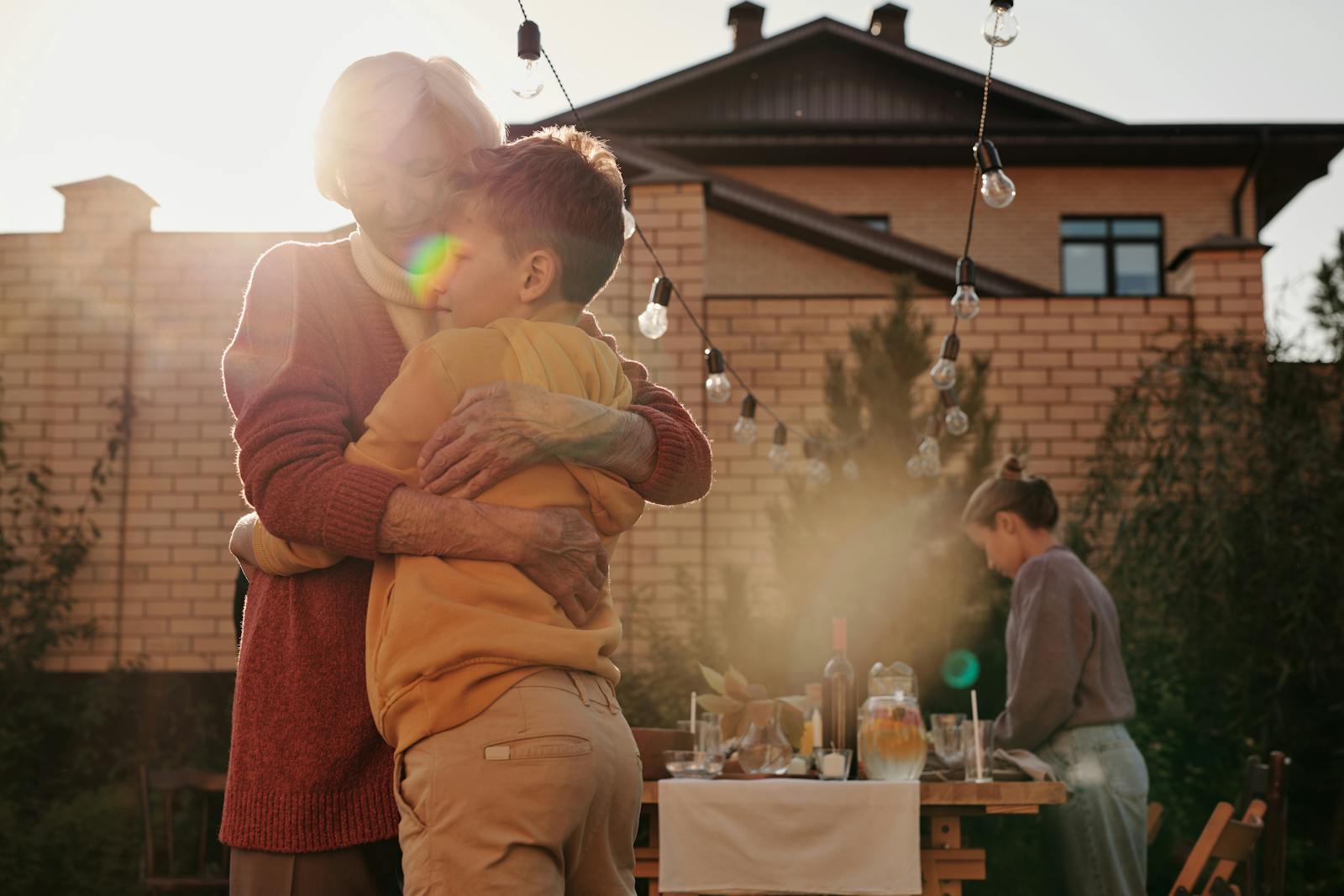 A grandmother hugging her grandson in a sunny backyard with an outdoor table setup.