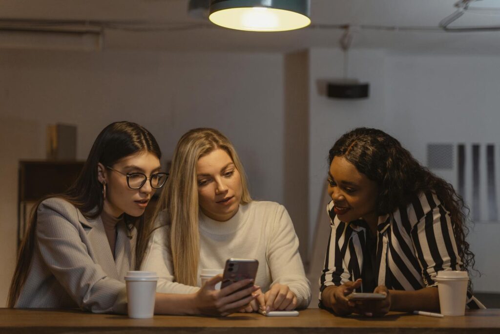 Three diverse women enjoying time together, interacting with smartphones at a table indoors.