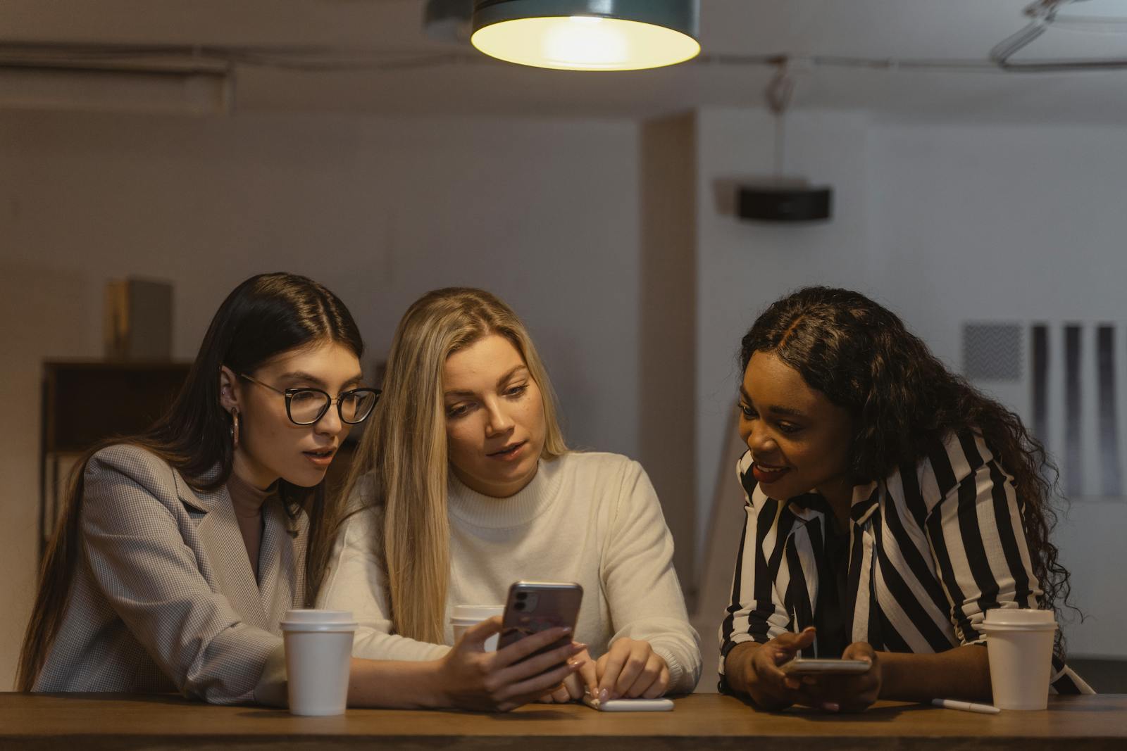 Three diverse women enjoying time together, interacting with smartphones at a table indoors.