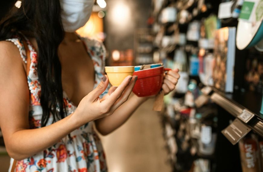 Woman in floral dress and face mask chooses ceramic mugs in store.