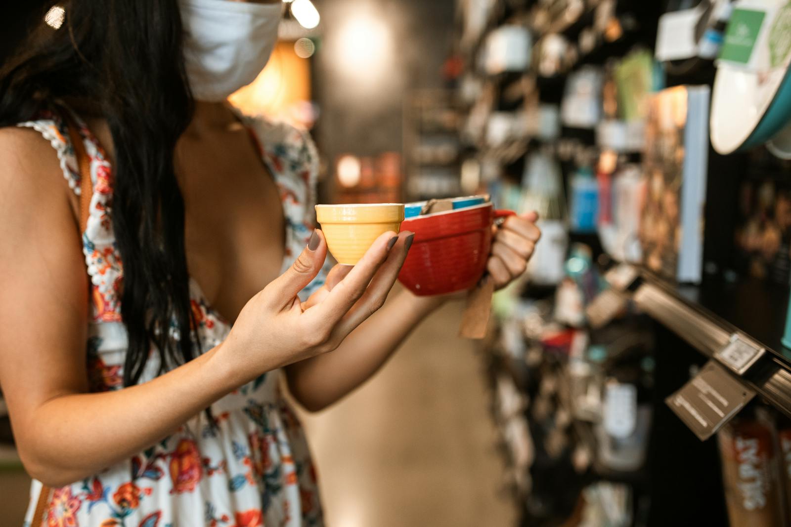 Woman in floral dress and face mask chooses ceramic mugs in store.