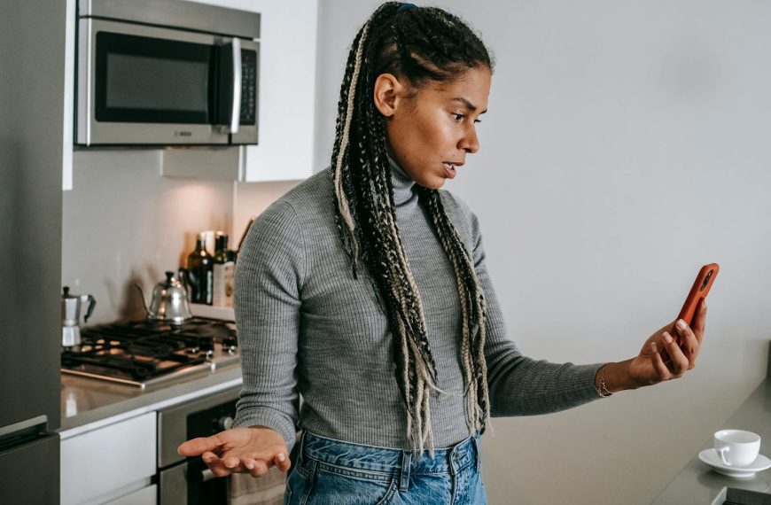 Serious young black lady with Afro braids in casual clothes gesticulating while having unpleasant conversation via video chat on smartphone in modern kitchen
