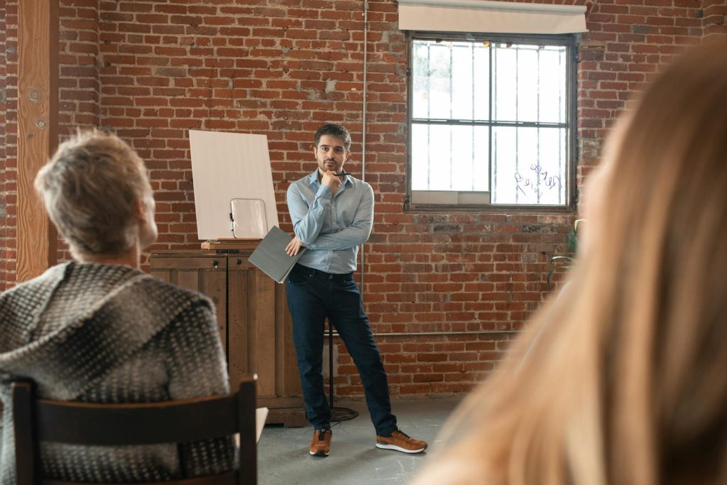 Teacher engaging adult students in a classroom discussion against a brick wall background.
