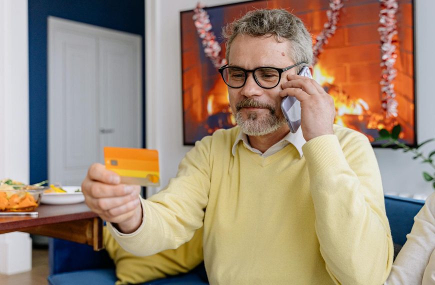 Elderly man using phone while holding a credit card indoors near a cozy fireplace.