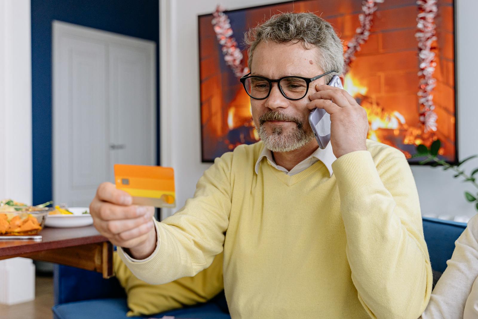 Elderly man using phone while holding a credit card indoors near a cozy fireplace.