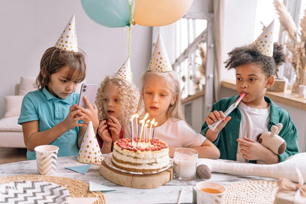 Group of children celebrating a birthday with cake, candles, and party hats indoors.