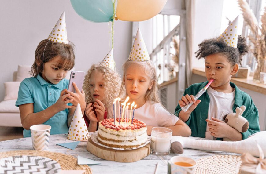 Group of children celebrating a birthday with cake, candles, and party hats indoors.