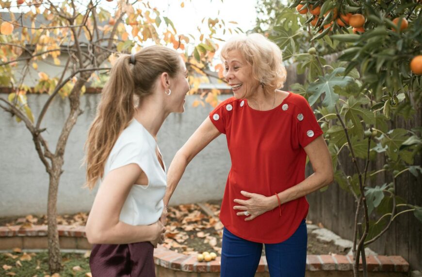 Two women laughing and talking under fruit trees in a garden, enjoying a sunny day.