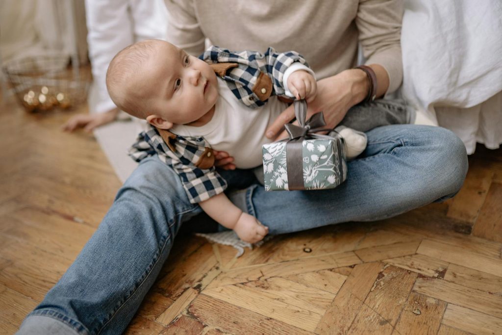 Cute baby holding a small gift while sitting on a parent's lap indoors.