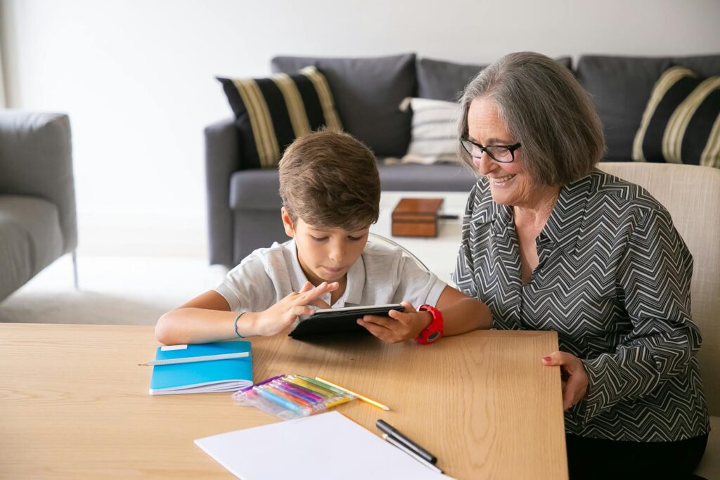 A senior woman smiles while helping her grandson with a tablet in a cozy living room.