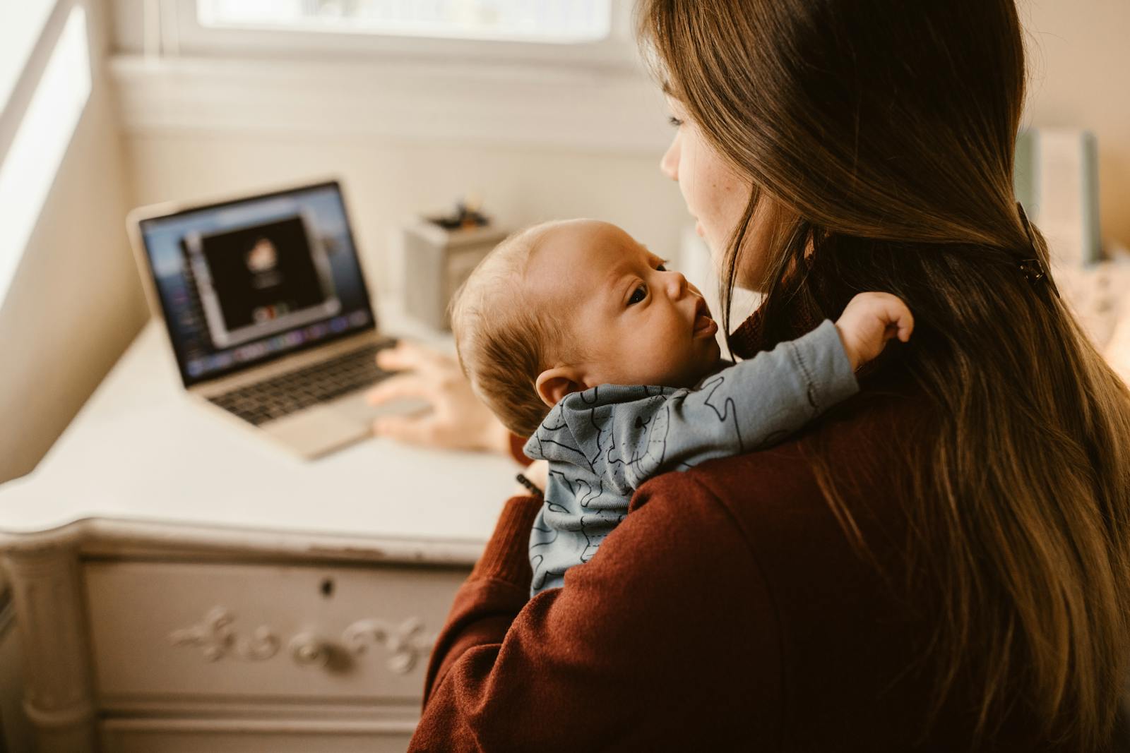 A mother multitasking at her desk, holding her baby while on a video call at home, showcasing modern parenthood.