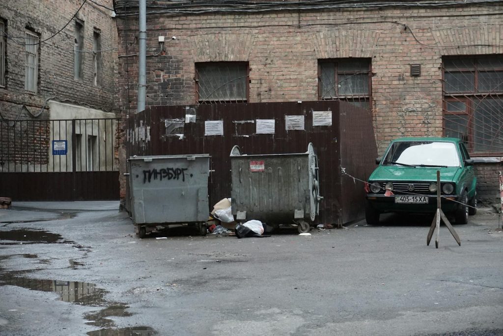 A gritty urban alley featuring garbage bins, scattered litter, and an old parked car.
