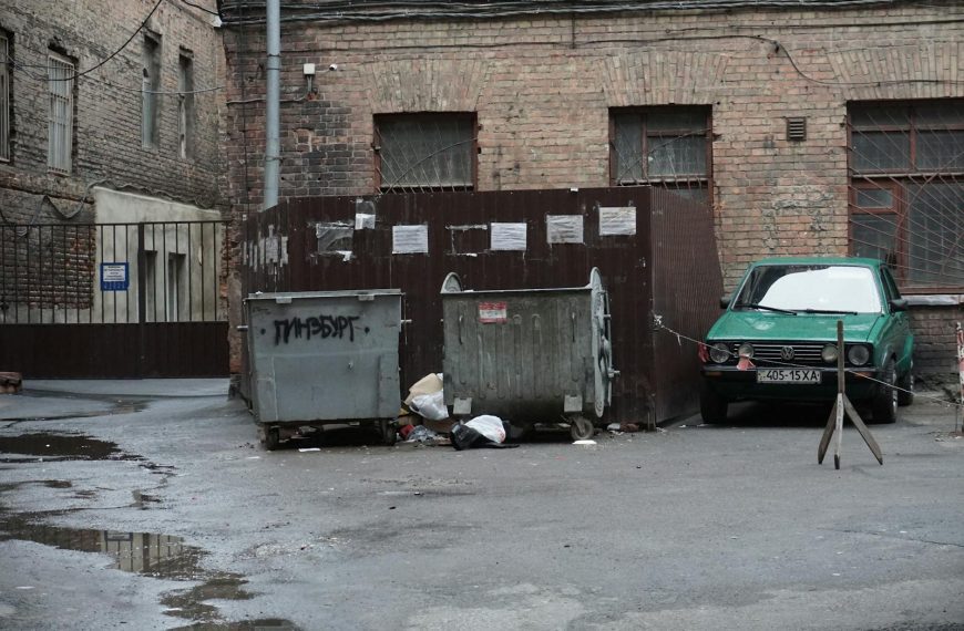 A gritty urban alley featuring garbage bins, scattered litter, and an old parked car.