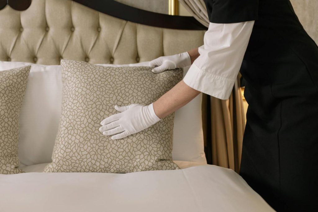 A housekeeper wearing gloves arranges pillows on a luxurious bed in a hotel room.