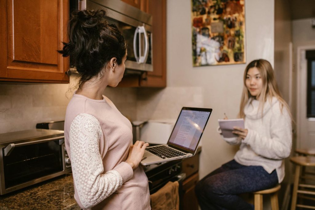 Two women collaborate on laptops and notes in a cozy home kitchen setting.