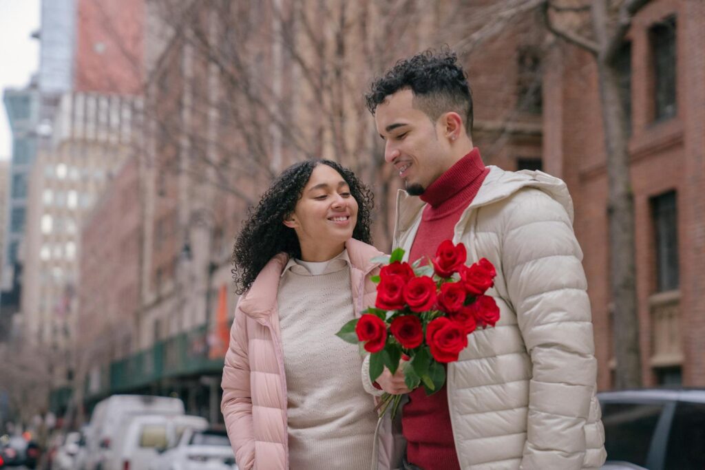 Young glad ethnic couple with bouquet of bright red roses smiling on urban street