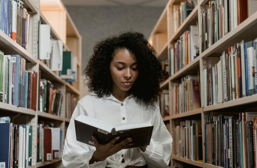African American woman with curly hair standing in a library reading a book.