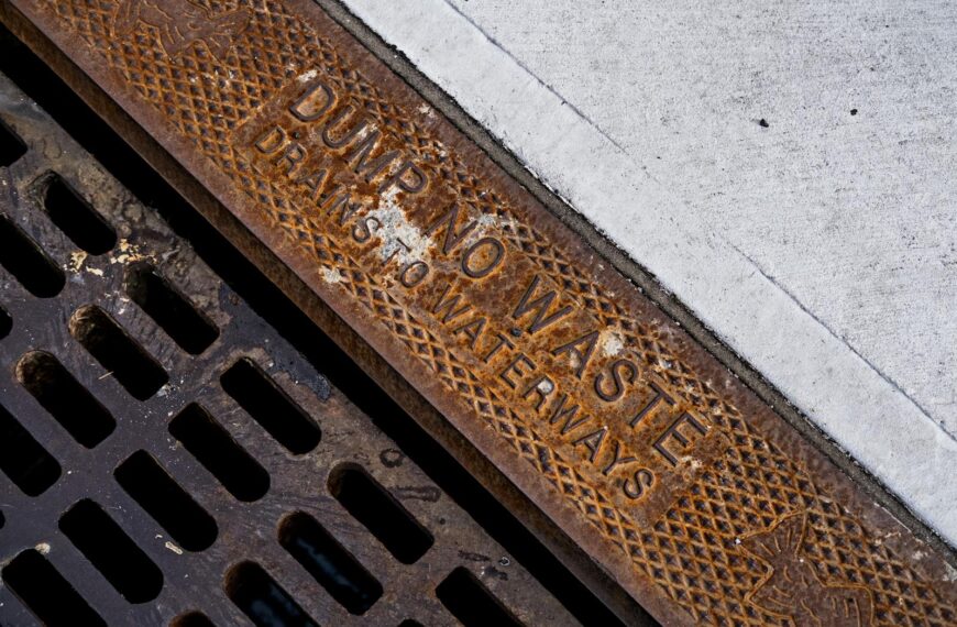 Close-up of a rusty drainage grate with environmental warning text on concrete sidewalk