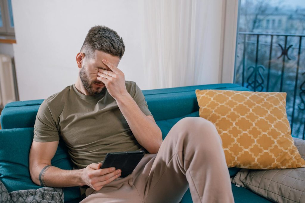 A man sits on a sofa holding a tablet, visibly stressed and covering his face with one hand.