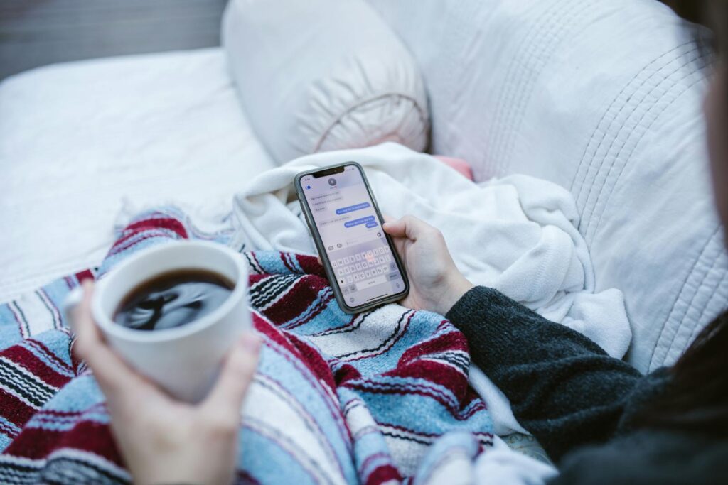 Person relaxing indoors with coffee and smartphone, texting in a cozy setting.