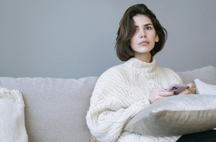 A thoughtful woman in a cozy sweater relaxing on a sofa while using her smartphone indoors.