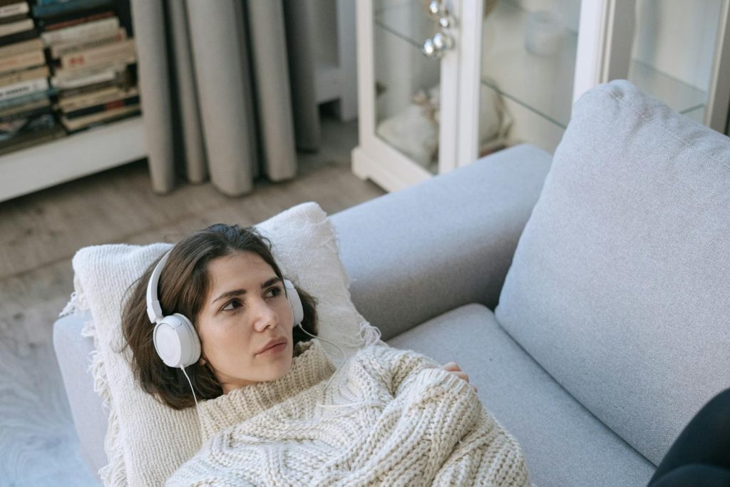 Young woman wearing headphones and knitted sweater, lying on a couch and relaxing indoors.