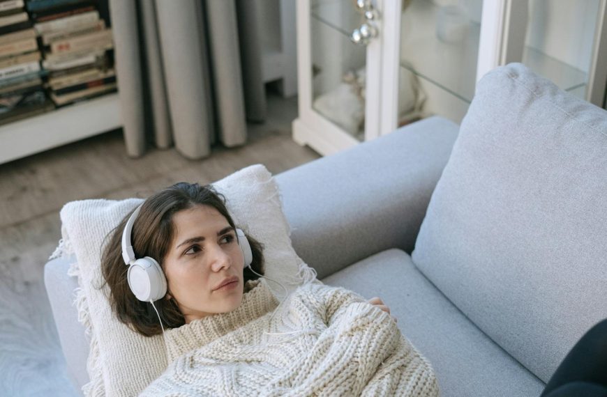 Young woman wearing headphones and knitted sweater, lying on a couch and relaxing indoors.