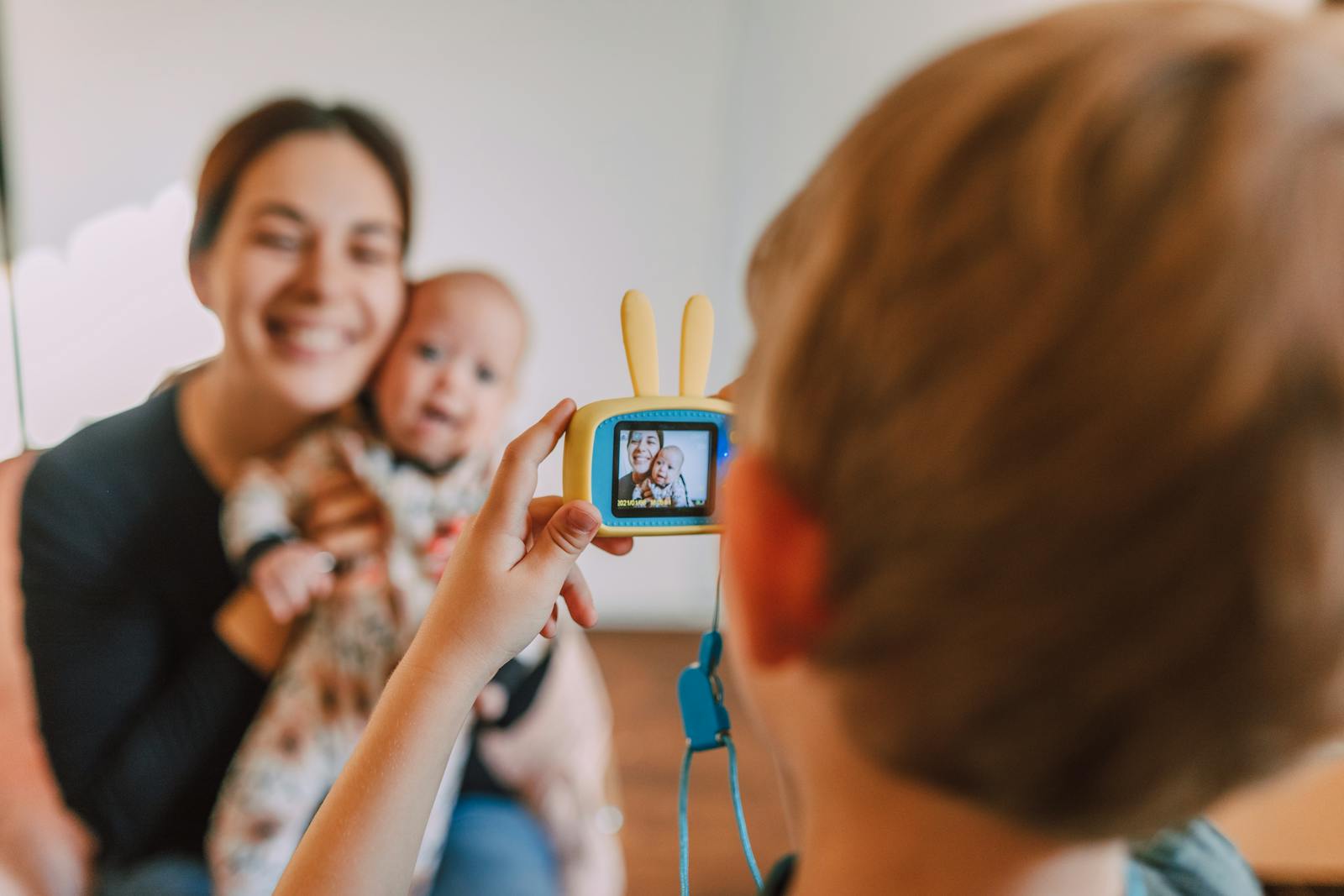 A child photographs their mother and baby sibling indoors, capturing a heartwarming family moment.