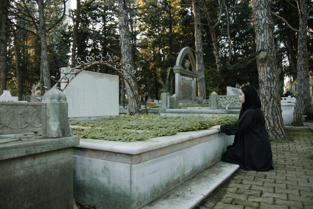 Side view of upset female in black outfit sitting on tiled walkway against tombstone in graveyard