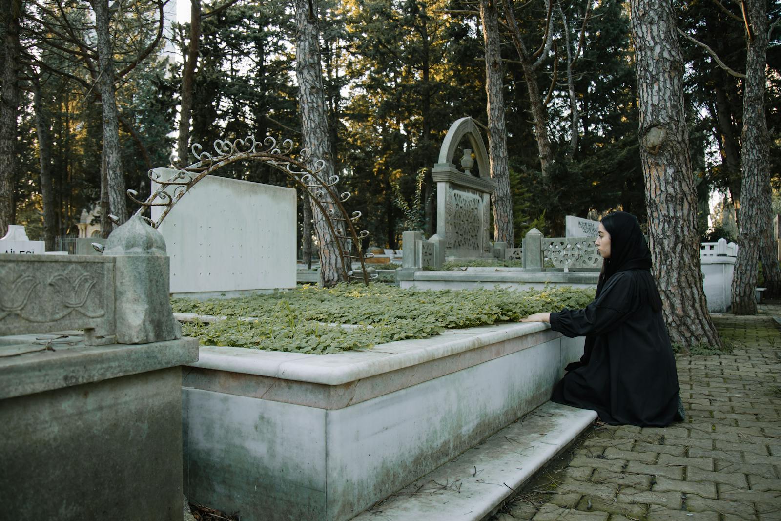 Side view of upset female in black outfit sitting on tiled walkway against tombstone in graveyard