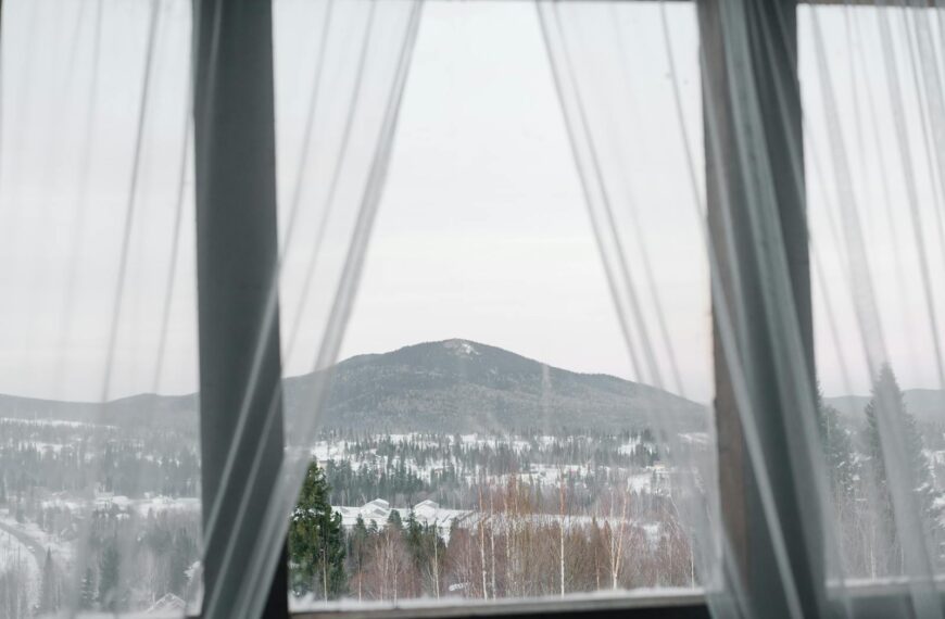 A serene winter landscape framed by sheer curtains, showcasing snow-covered mountains and forests.
