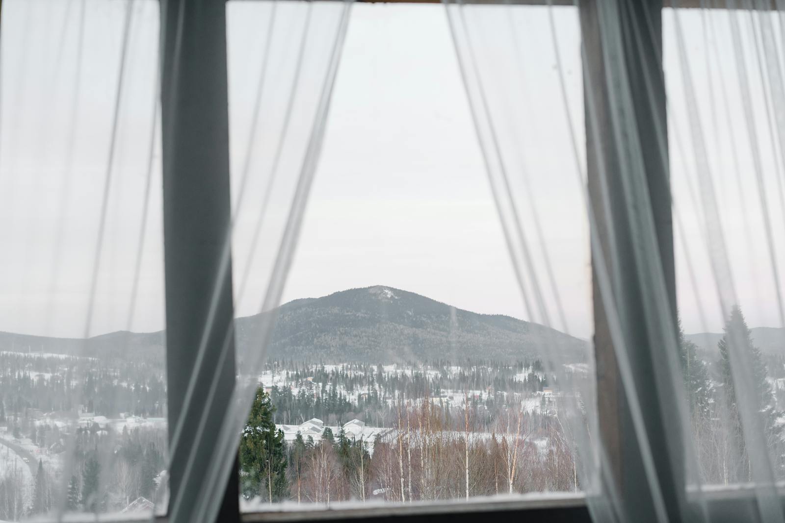 A serene winter landscape framed by sheer curtains, showcasing snow-covered mountains and forests.