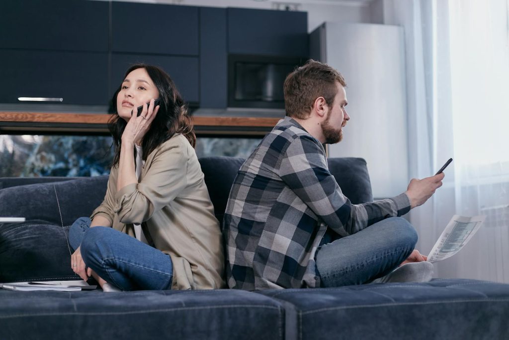 A couple sitting back-to-back on a sofa, expressing financial tension while using phones.