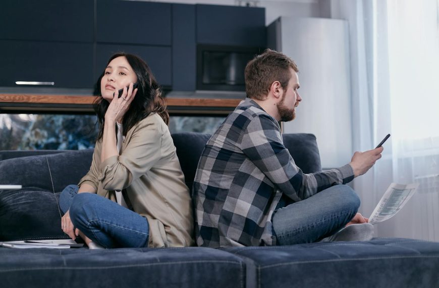 A couple sitting back-to-back on a sofa, expressing financial tension while using phones.