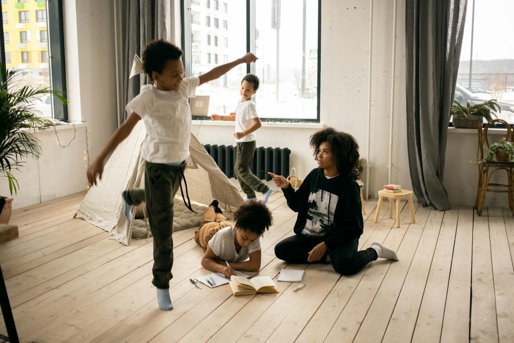 Full body of African American mother sitting on wooden floor near daughter writing in copybook in light room with playing boys