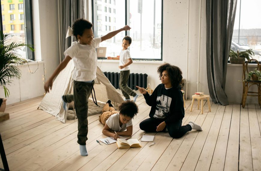 Full body of African American mother sitting on wooden floor near daughter writing in copybook in light room with playing boys