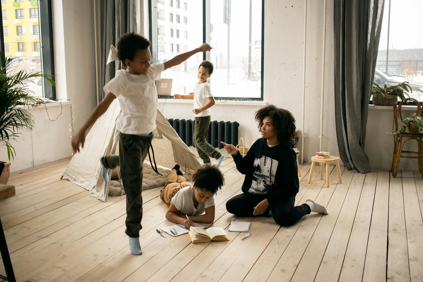 Full body of African American mother sitting on wooden floor near daughter writing in copybook in light room with playing boys