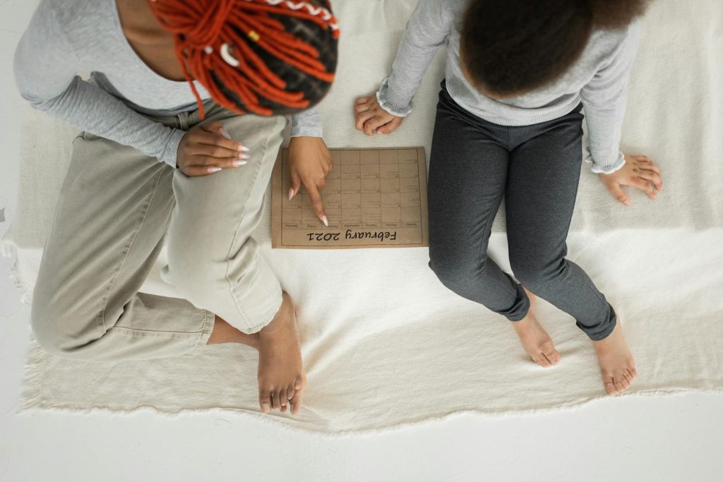 From above of crop anonymous African American female in casual clothes sitting on bed with girl and reading textbook