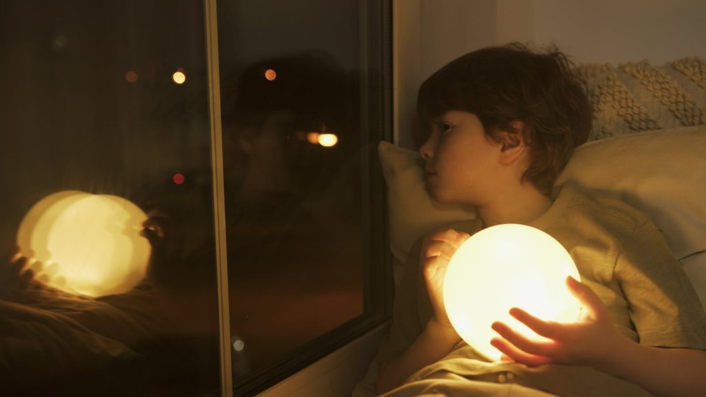 A young boy holding a glowing orb, looking out a window at night.