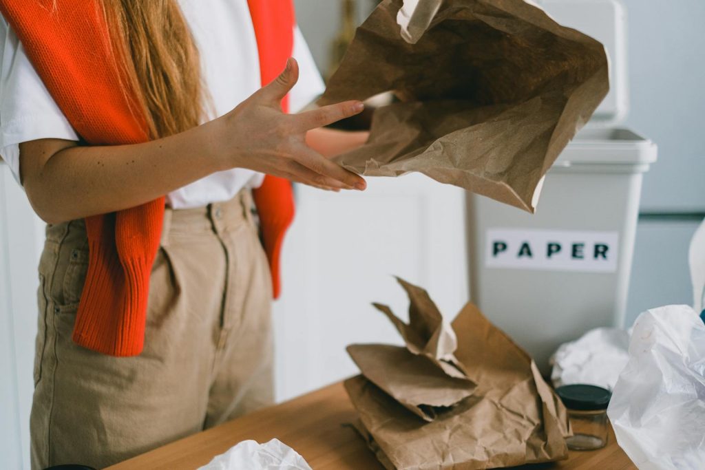 Crop anonymous female standing at table and sorting paper trash from plastic packages and glass