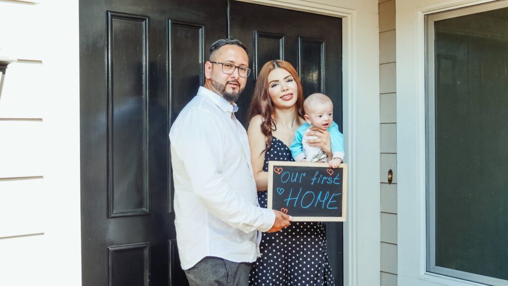 Young family with baby holding a sign 'Our First Home' at doorstep, symbolizing new beginnings.