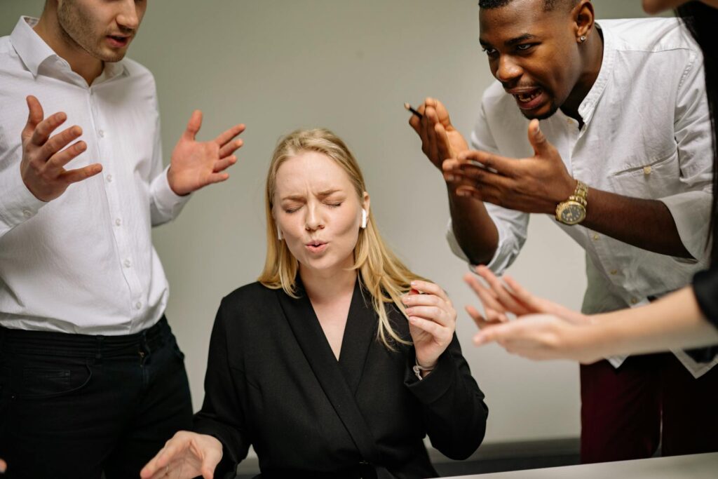 A stressed woman in an office surrounded by arguing coworkers highlighting workplace tension.