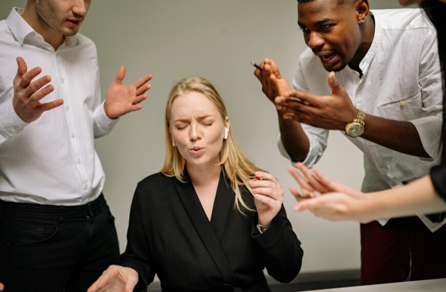 A stressed woman in an office surrounded by arguing coworkers highlighting workplace tension.