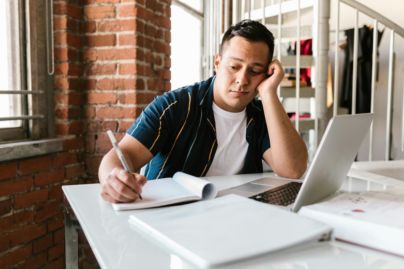 A student looks bored while studying with a laptop and notebook on a table.