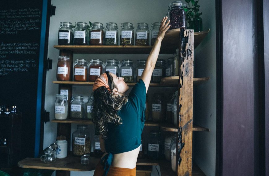 Woman reaching for a jar on a shelf in an eco-friendly shop with wooden decor and glass containers.